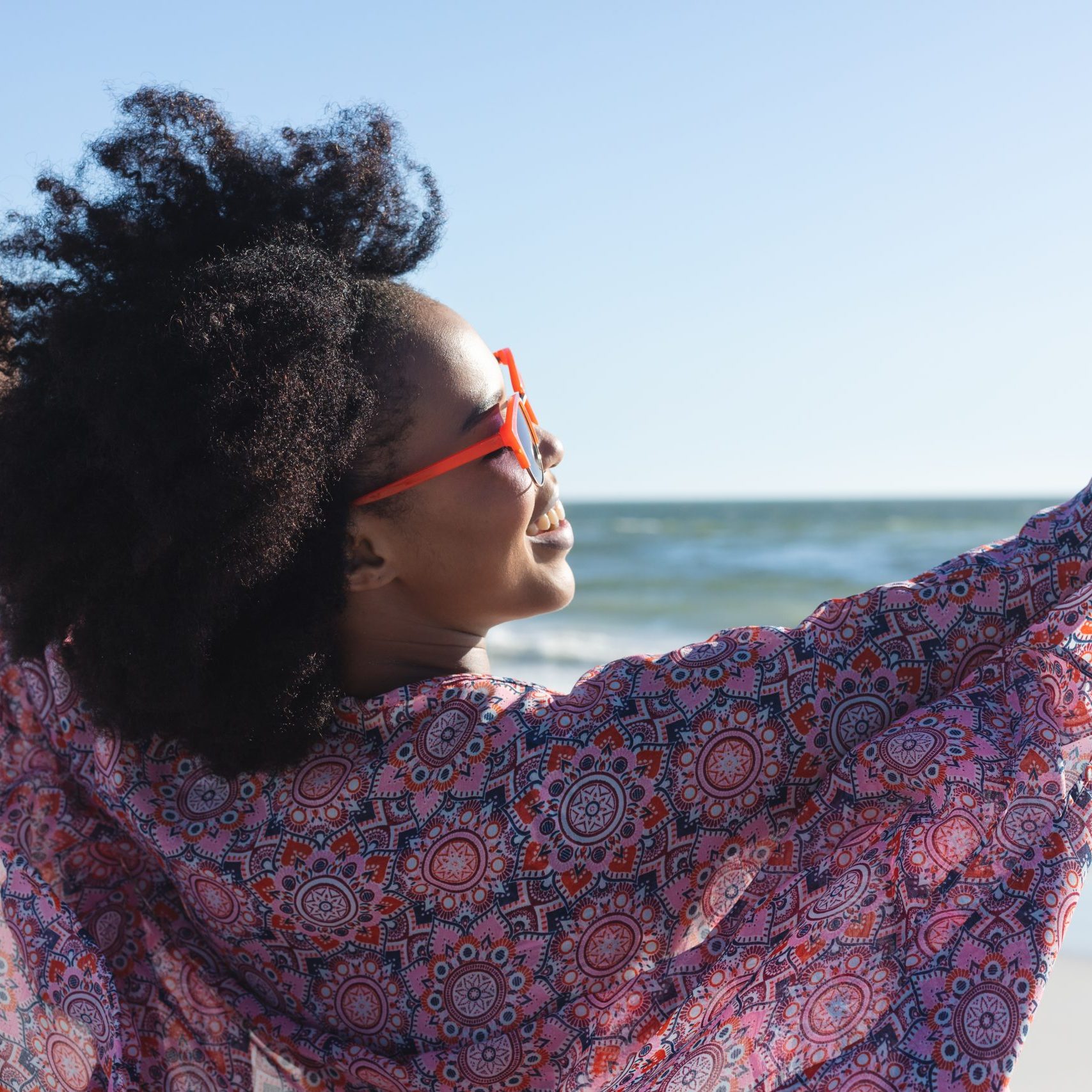 Happy african american woman in sunglasses smiling with arms outstretched on sunny beach by sea. Summer, wellbeing, freedom, relaxation and vacation, unaltered.